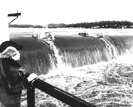 Water flowing over a dam; young boy in winter clothing standing at handrail in lower left corner